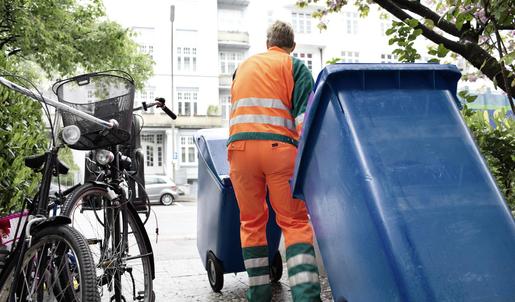 Ein Müllwerker in Warnkleidung schiebt eine blaue Mülltonne vor sich her und zieht eine blaue Mülltonne hinter sich her.